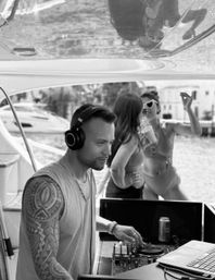 Male DJ wearing headphones and a tribal arm tattoo mixing on a deck-mounted controller aboard a yacht, with two women in swimsuits dancing and sipping drinks in a waterfront boat-party scene.
