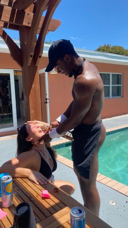 Playful poolside scene: shirtless man in a black cap and striped apron feeding a laughing woman in a black bikini beside a backyard pool under a wooden pergola on a sunny summer day, drinks on a wooden table.