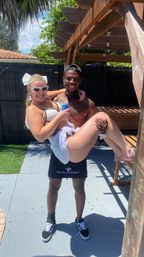 Smiling shirtless man in a cap carries a laughing woman in a white bikini and cat-eye sunglasses, who holds a canned drink under a wooden pergola on a sunny backyard patio.