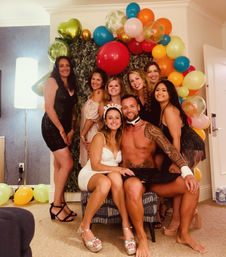 Bachelorette party group photo in a decorated hotel suite: bride-to-be in white dress and “BRIDE” headband sits on a shirtless entertainer’s lap, friends in colorful party dresses pose under a vibrant balloon arch.