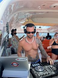 Shirtless tattooed DJ wearing headphones and sunglasses mixing on a yacht deck with a laptop sticker reading I Love London, a DJ controller in the foreground and partygoers relaxing in the background.