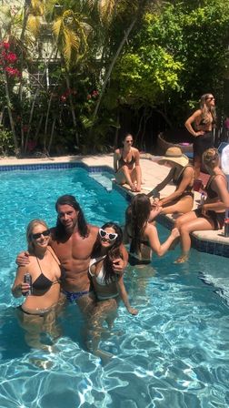 Sunny summer pool party in a residential backyard: friends in bikinis and swim trunks posing in clear blue pool water by a tiled deck with palm trees and tropical landscaping.