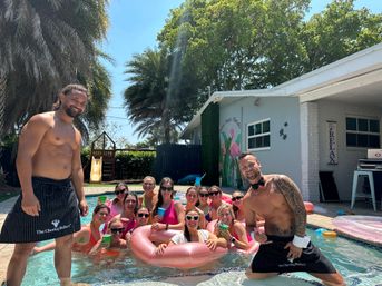 Smiling group of women at a sunny backyard pool party on a large pink inflatable, flanked by two shirtless attendants, palm trees and a house with a flamingo mural in the background.