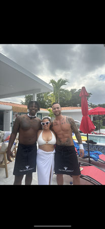 Poolside party: woman in white bikini and heart-shaped sunglasses stands between two shirtless servers wearing black aprons by a tropical backyard pool with red umbrellas and palm trees under a cloudy sky.