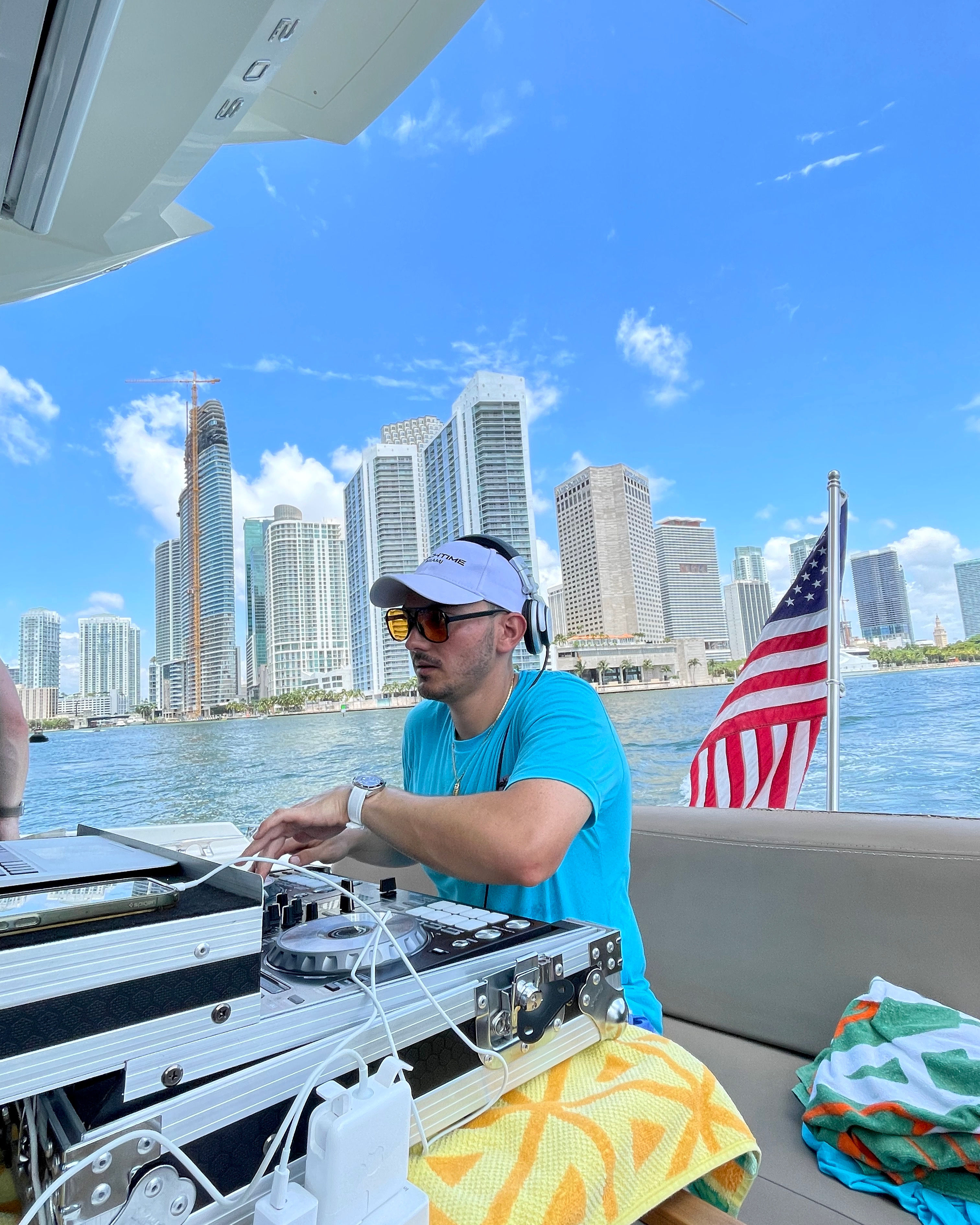 DJ mixing on a boat during a Miami waterfront boat party, laptop and turntables on deck, American flag and downtown skyline under a bright blue sky.