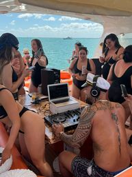 Yacht party scene: tattooed DJ in a captain's hat spins on a laptop and mixer as women in black swimsuits dance and sip drinks on a sunny ocean day with a boat on the horizon.