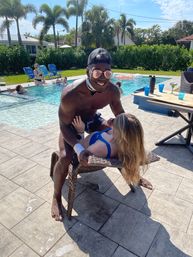 Playful summer pool party scene: smiling man in sunglasses and cap leaning over a woman in a blue swimsuit on a wicker chair beside a sunny backyard pool with palm trees, floats, and friends lounging nearby.