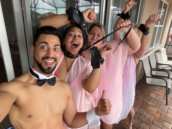 Playful adult group selfie on a tiled backyard patio: shirtless man in a bow tie and three women in pink robes laughing while holding leather wrist restraints and a leash.