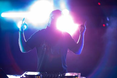 Silhouetted DJ with raised arms behind illuminated turntables, vibrant purple-blue stage lights and lens flare at a live club show