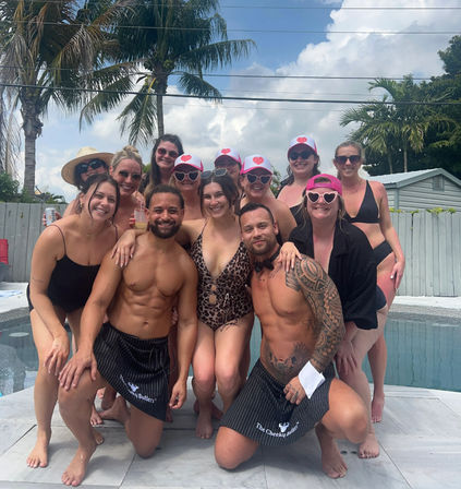 Smiling group of women in swimsuits and two shirtless male entertainers posing by a backyard pool with palm trees and sunny sky, pink caps and heart-shaped sunglasses for a tropical pool party vibe