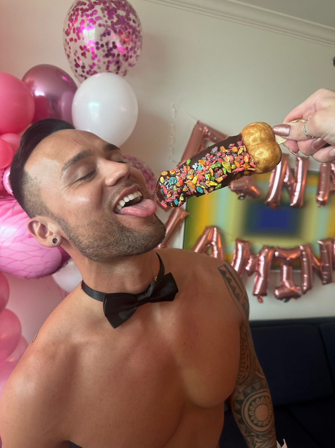 Shirtless partygoer in a black bow tie playfully licking a chocolate-coated dessert pop topped with colorful cereal, with pink and white balloons and rose-gold letter balloons in the background at an indoor birthday celebration.