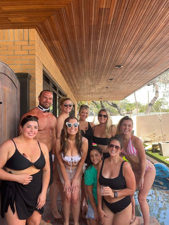 Cheerful group of adults in swimwear posing on a shaded backyard poolside patio under a wood-paneled overhang on a sunny day.