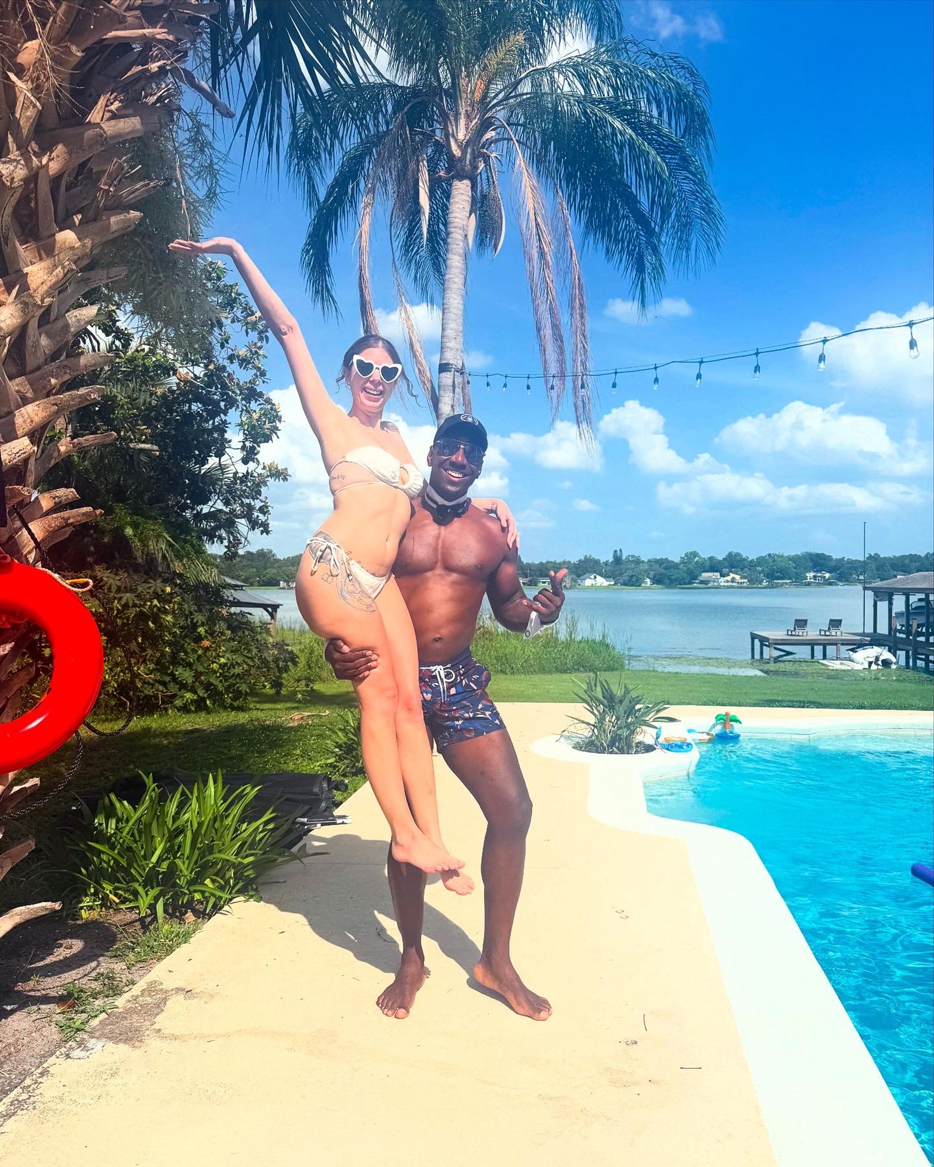 Smiling woman in a white bikini and heart-shaped sunglasses lifted by a shirtless man in swim trunks at a sunny lakeside backyard pool, framed by a palm tree, string lights, dock and blue sky.