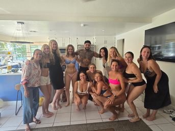 Smiling group of adults in swimsuits posing together in a bright vacation-style kitchen with a backyard pool visible through the windows.