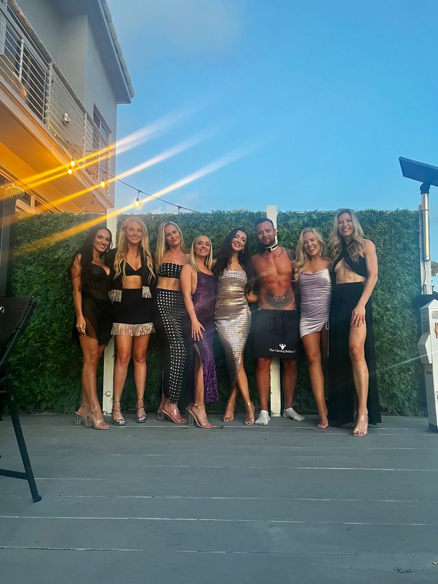 Group of eight people posing on an outdoor deck at dusk, women in glamorous sparkly dresses and heels and a man in a bow tie and apron, string lights and a green hedge backdrop under a clear blue sky.