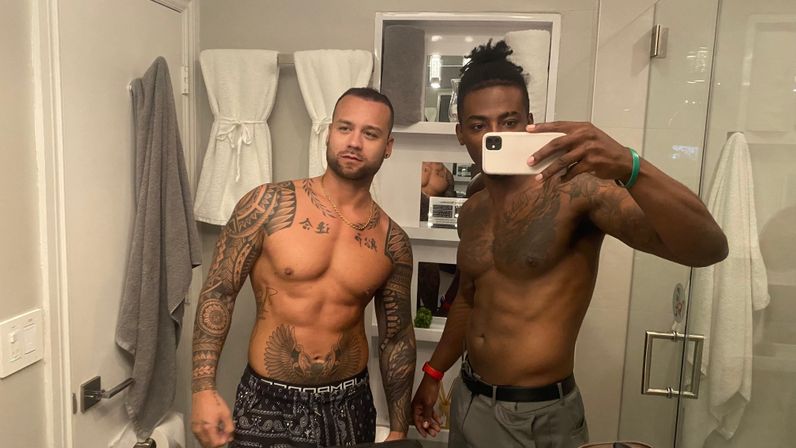 Bathroom mirror selfie of two shirtless men with tattoos posing in a modern home bathroom with glass shower and white towels