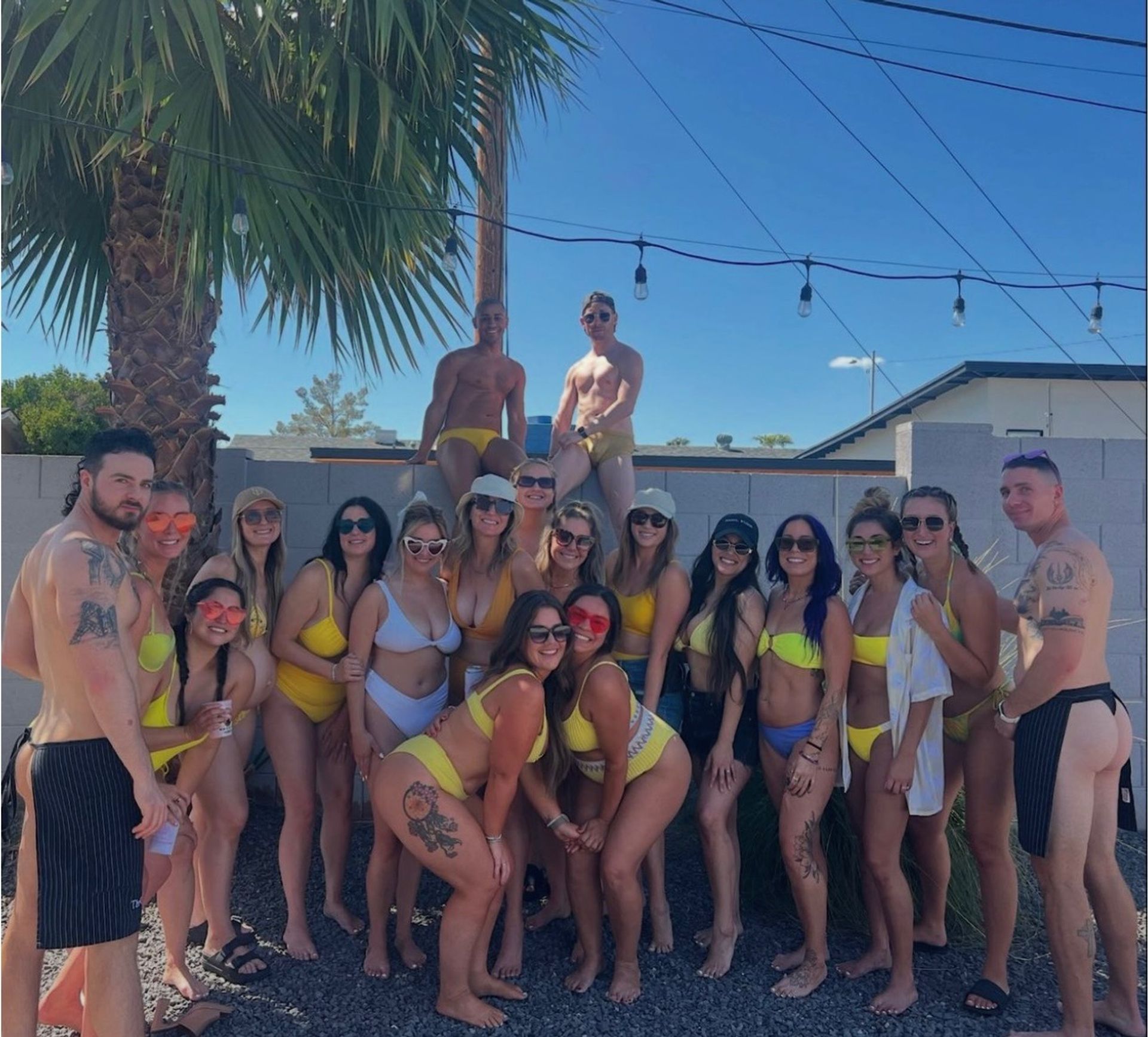 Large group of young adults in yellow and white swimsuits posing at a sunny backyard pool party under a palm tree and string lights against a bright blue sky