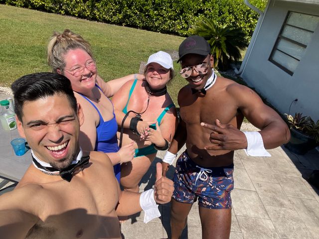 Sunny backyard pool-party selfie of four friends on a patio: two shirtless men wearing bow ties and cuffs and two women in colorful swimsuits smiling and posing.