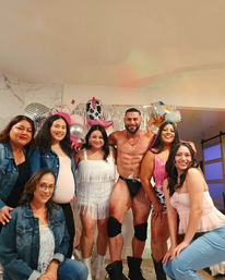 Smiling group of women posing in a living room with a muscular male entertainer in black dancewear, silver fringe backdrop and pink metallic balloons including a cowgirl boot balloon — lively indoor bachelorette-style party.