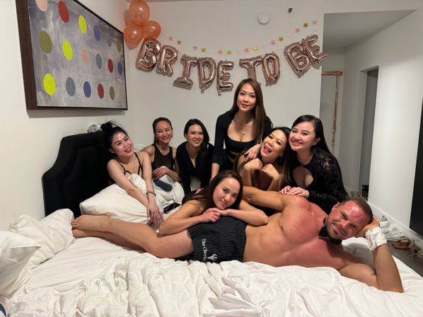 Seven women posing on a bed at a bachelorette party with a metallic "Bride to Be" balloon banner and a shirtless male entertainer reclining at the foot of the bed in a decorated bedroom.