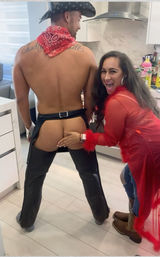 Playful home kitchen costume party: woman in red sheer robe laughing as she playfully touches the exposed butt of a shirtless man wearing a cowboy hat and leather chaps.