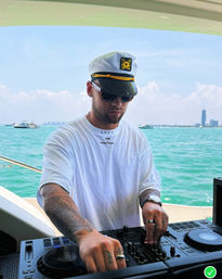 DJ in a white captain's hat and sunglasses mixing on a yacht deck, tattoos and rings visible, turquoise ocean and distant coastal skyline under a sunny blue sky