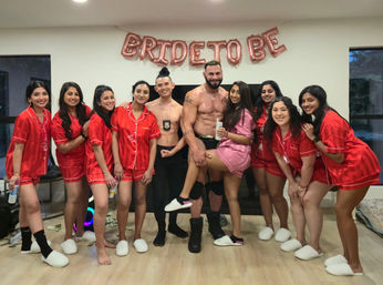 Bachelorette party in a living room with rose-gold 'BRIDE TO BE' balloons: bride-to-be in a pink satin robe being held by a shirtless male entertainer, surrounded by smiling friends in matching red satin pajama sets and white slippers.
