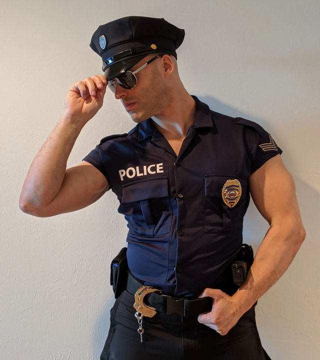 Playful portrait of a person in a navy police uniform and cap tipping sunglasses, handcuffs on belt, posed against a plain indoor wall.