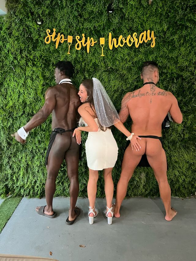 Bachelorette party photo booth: bride in a short white dress and veil poses playfully between two shirtless male entertainers in black aprons, each showing their backsides in front of a green foliage wall with a gold “sip sip hooray” sign.