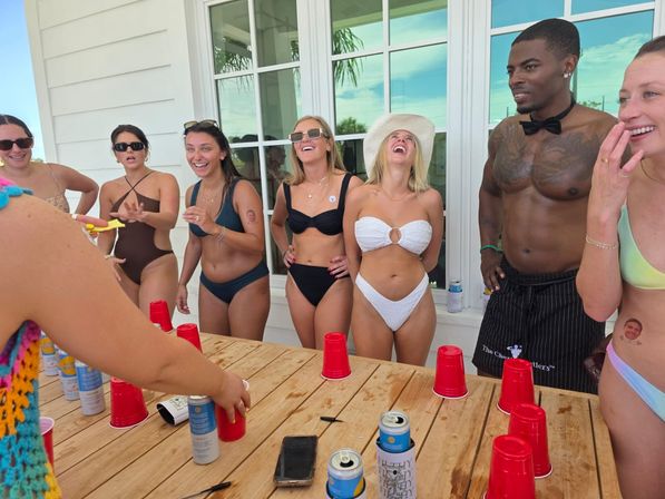Adults in swimwear laughing on a beach-house patio, gathered around a wooden table playing a red-cup party game with drinks and phones; woman in white bikini and sun hat laughs while a shirtless man in a bow tie stands nearby.