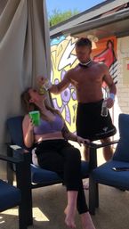 Sunny poolside cabana scene: shirtless attendant playfully feeds green grapes to a seated woman with a green cup, colorful graffiti mural in the background.
