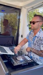 Tattooed DJ in sunglasses and patterned shirt mixing on a Pioneer DJ controller and laptop aboard a boat with sunny leafy shoreline in the background