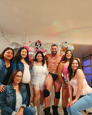 Seven women smiling and posing with a muscular shirtless entertainer in a black thong and knee pads at an indoor party with pink balloons, disco-style decor and a silver fringe backdrop.