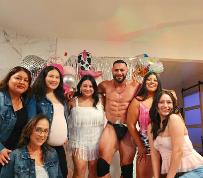 Seven women smiling and posing with a muscular shirtless entertainer in a black thong and knee pads at an indoor party with pink balloons, disco-style decor and a silver fringe backdrop.