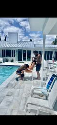 Sunny poolside scene: woman in orange dress sits on a white chair while two shirtless men playfully attend to her on a modern white backyard patio with wicker seating and a blue pool.