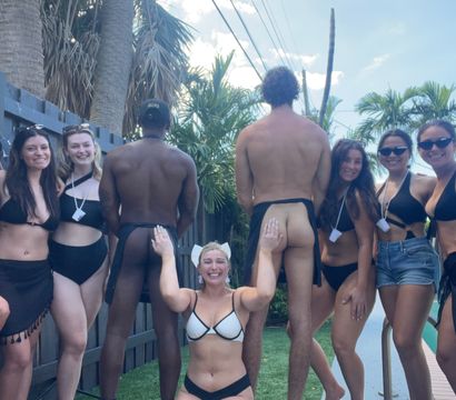 Sunny backyard pool party: group of friends in black swimwear posing by a pool, with two men facing away in cheeky swim bottoms and a woman kneeling playfully between them, palm trees and fence in the background.