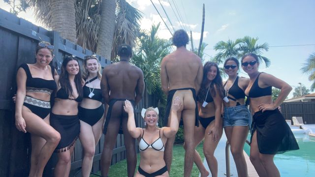 Sunny backyard pool party: group of friends in black swimwear posing by a pool, with two men facing away in cheeky swim bottoms and a woman kneeling playfully between them, palm trees and fence in the background.
