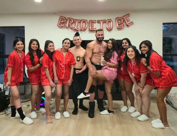 Bachelorette party in a living room: bride-to-be in pink and friends in matching red satin pajamas posing and smiling with two shirtless men under rose-gold 'BRIDE TO BE' balloons.