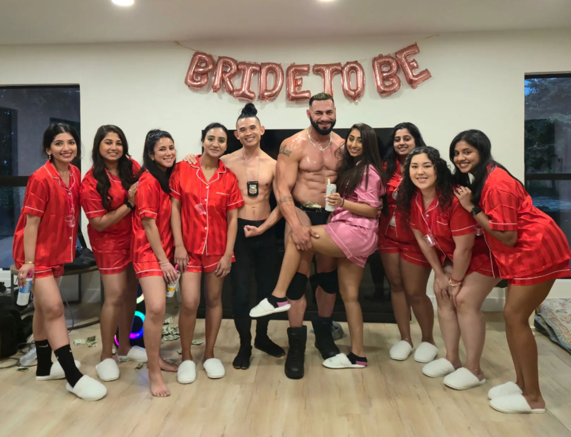 Bachelorette party in a living room: bride-to-be in pink and friends in matching red satin pajamas posing and smiling with two shirtless men under rose-gold 'BRIDE TO BE' balloons.