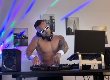 Shirtless, tattooed DJ in white sunglasses and headphones mixing on a controller with laptop and speakers, colorful disco lights and black-and-white cityscape prints on the wall.