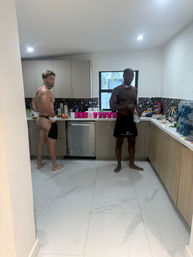 Two shirtless men in playful bow ties and server aprons prepping drinks in a modern kitchen with marble floor, terrazzo backsplash, pink party cups, snacks and a window showing palm trees.