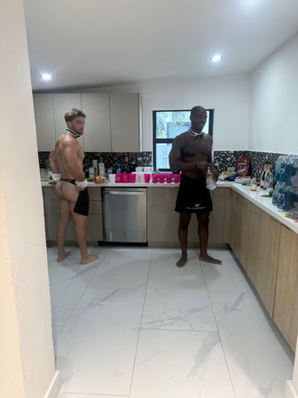 Two shirtless men in playful bow ties and server aprons prepping drinks in a modern kitchen with marble floor, terrazzo backsplash, pink party cups, snacks and a window showing palm trees.