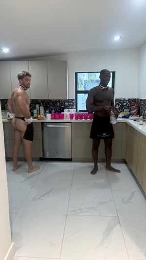 Two shirtless men in bow ties and black aprons preparing drinks in a modern home kitchen with pink party cups, snacks and bottled water on the counter and marble-tiled floor.