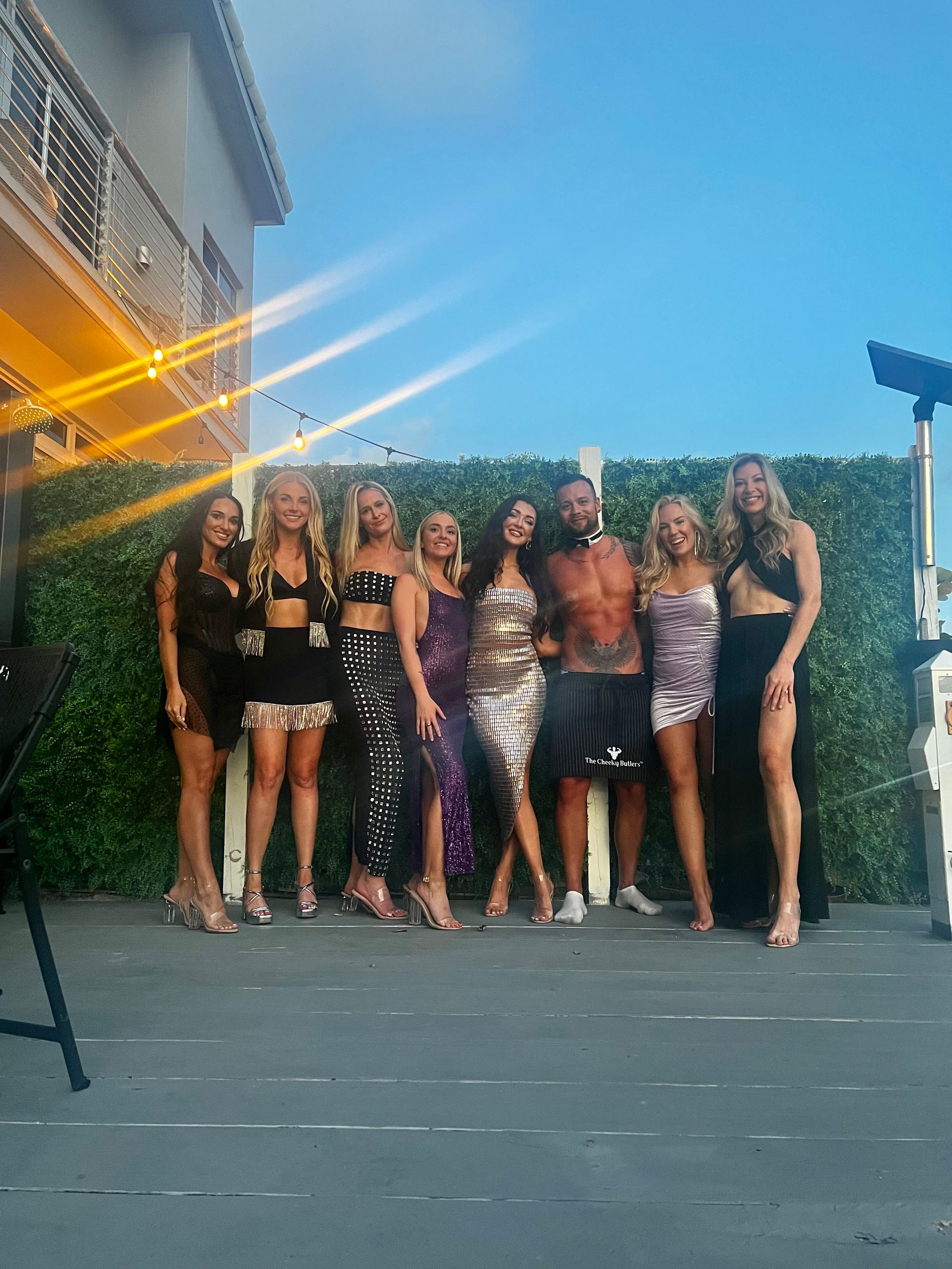 Eight adults in sparkly party outfits posing on a backyard patio at dusk under string lights and a clear blue sky.