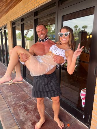 Smiling shirtless man in a bow tie and pinstripe skirt lifts a woman in a cherry-print bikini, heart-shaped sunglasses and sheer pearl skirt on a sunny poolside patio with palm trees.