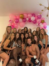 Group of smiling women with two shirtless male dancers in bow ties posing under a pink-and-gold balloon arch at an indoor bachelorette party