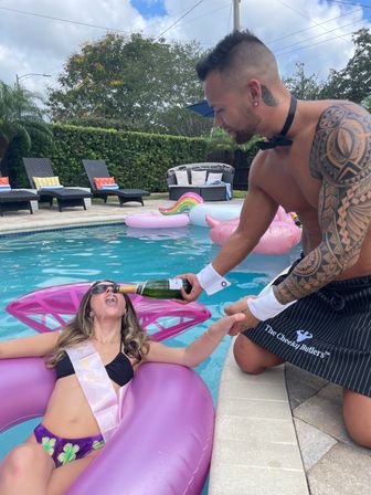 Shirtless tattooed man wearing a bow tie pours champagne into the mouth of a woman in a sash who’s lounging on a pink inflatable in a backyard pool during a sunny pool party.