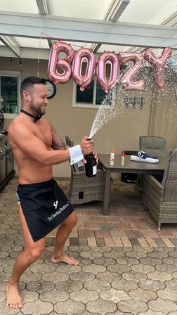 Shirtless person in a black apron and cuff playfully pops a champagne bottle, spraying bubbly under rose-gold foil balloons above a backyard patio with wicker chairs and a dining table.