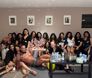 Indoor living room party: a group of smiling women gathered on a couch and floor around a shirtless male entertainer posing in front of a coffee table with drinks, snacks and cash.