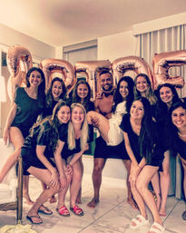 Bachelorette party: smiling women and a man pose indoors as the bride-to-be in white is lifted in front of large rose-gold "BRIDE" balloons.